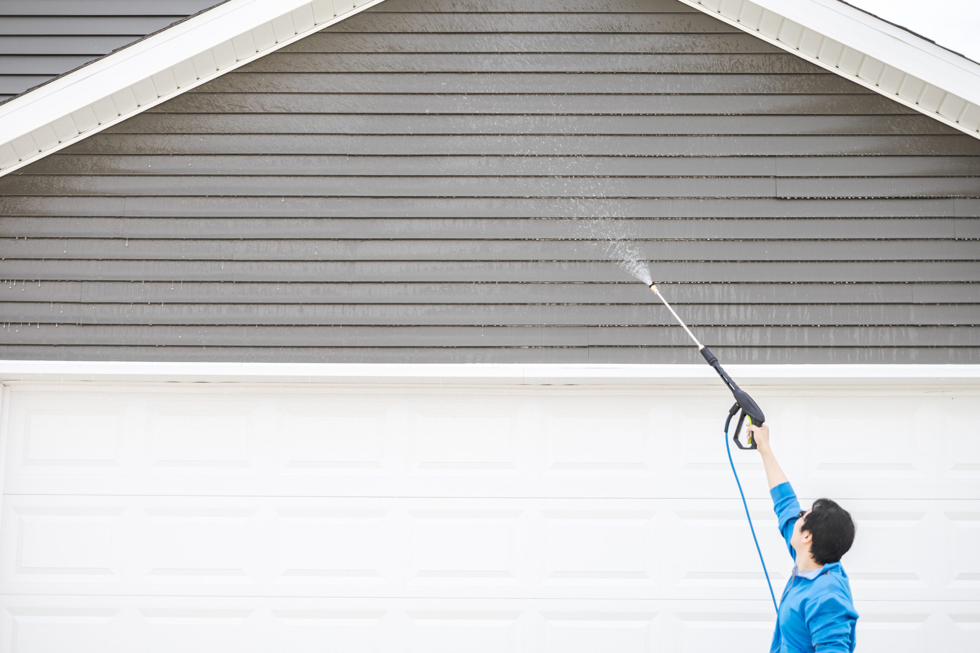 Man in blue jacket clean dusk and dirt from exterior siding under the roof at front door garage by high pressure nozzle spray with water soap cleaner, weekend chore. Washer machine of cleansing house. Man in blue jacket clean dusk and dirt from exterior siding under the roof at front door garage by high pressure nozzle spray with water soap cleaner, weekend chore. Washer machine of cleansing house.